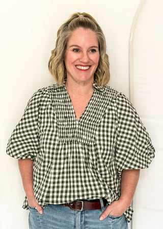 A smiling woman with blonde hair stands against a white background wearing a green and white gingham wrap-style blouse with puff sleeves and blue jeans.