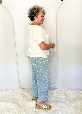 A woman stands in profile wearing a white short-sleeve top and light blue pants with white polka dots, posed in front of three large white arch backdrops on a white faux fur rug over a wood floor.