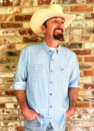 A man wearing a light blue and white gingham button-down shirt, a cream cowboy hat, and jeans, standing against a rustic brick wall.