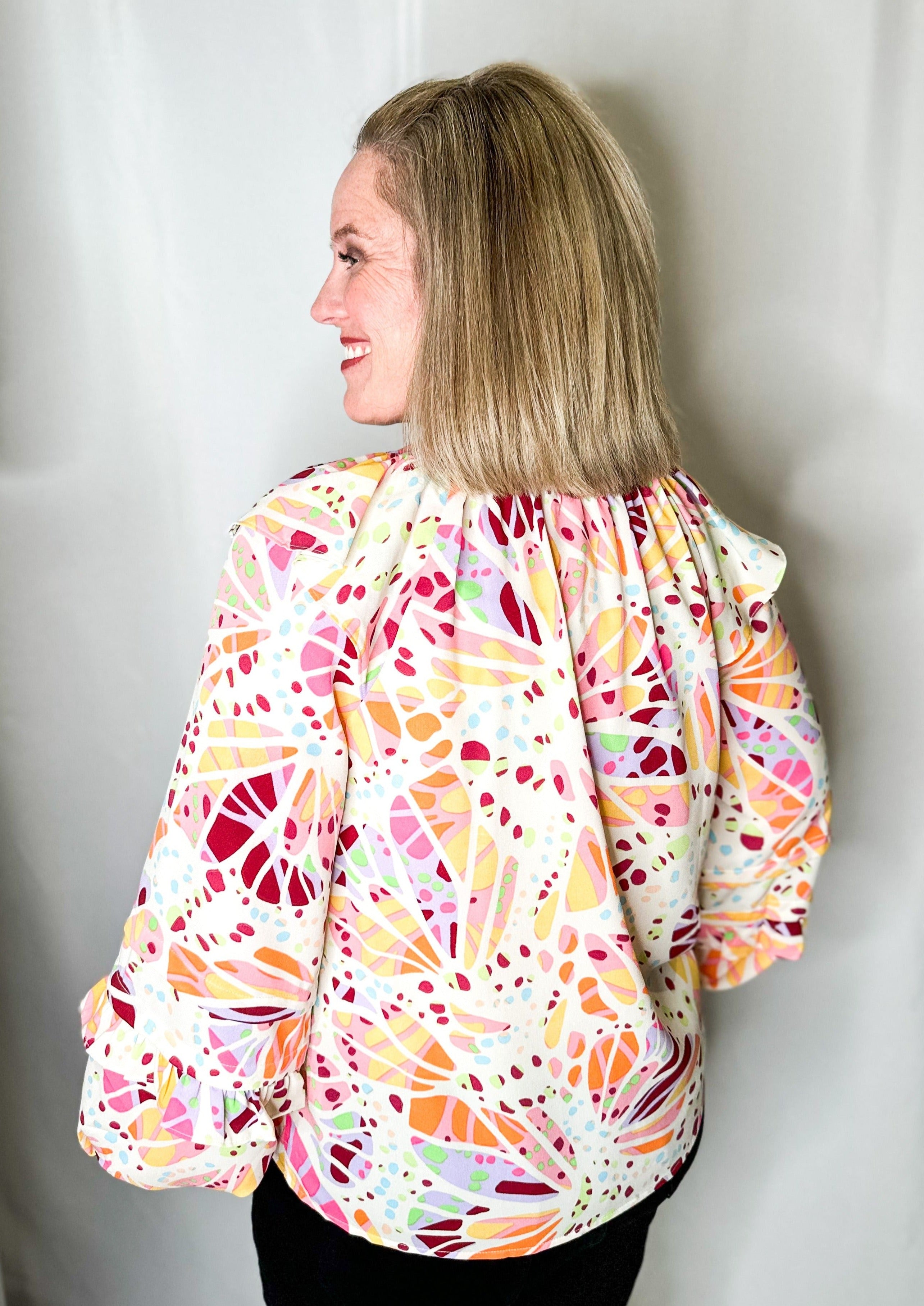 Back view of blouse. White long sleeve blouse with ruffle detail at the top of the sleeve near the shoulder and another small ruffle 3/4 down the sleeve. It has a higher neckline and multicolored abstract print all over.