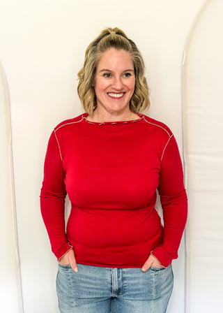 A smiling blonde woman wearing a bright red long-sleeve crewneck sweater and light blue jeans, standing against a white arched studio backdrop.