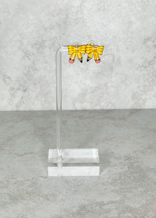 Yellow pencil bow-shaped earrings on a clear stand against a gray marble background