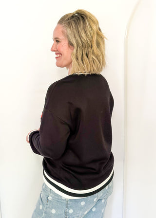 A woman with blonde hair tied back is shown from the back wearing a dark brown crewneck sweatshirt with black and white striped ribbed trim, paired with light blue polka dot jeans, against a white wall.