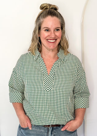 A smiling woman with blonde hair in a bun wears a green and white gingham button-down shirt with rolled-up sleeves, paired with blue jeans, standing against a white arched wall.