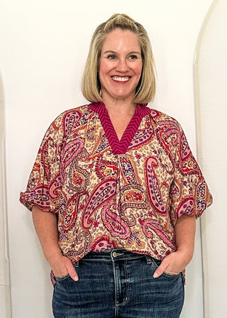 A woman stands in front of three large white arched panels in an indoor room, wearing a short-sleeve paisley blouse with red, beige, and green tones and blue jeans.