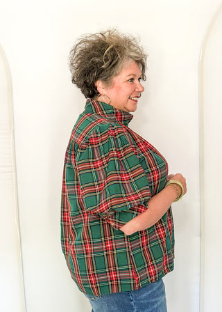 A woman with short curly gray hair is shown from the side, wearing a green and red plaid mock-neck shirt and blue jeans, standing against a white arched backdrop.