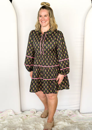 Long sleeve black knee-length dress. Pink and green floral design. Tiered with pink piping detail. Buttons on top.