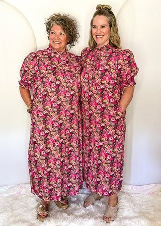 Two women standing side-by-side wearing matching high-neck midi dresses with a vibrant pink, red, and green floral print, posed in front of a white circular backdrop.