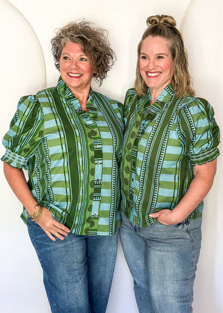 Two women standing side-by-side wearing matching green and light blue patterned button-up shirts and blue jeans, posing against a white arched backdrop.