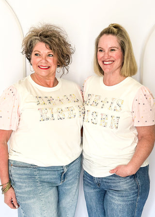 Off white short sleeve shirt with colorful embellished text and pink statement sleeves with decorative pearl studs.