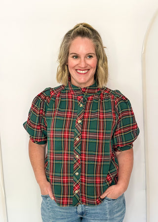 A smiling woman with blonde hair stands against a white arched backdrop, wearing a green, red, and yellow plaid short-sleeve button-up shirt with light blue jeans.