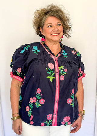 Navy floral short sleeve blouse with pink, green, yellow, and blue flowers, featuring puff sleeves, ruffled neckline, button front with pink trim, and a relaxed fit