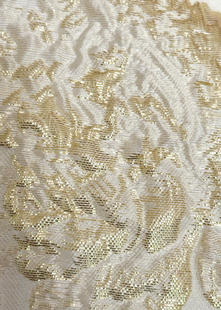 A close-up of a textured fabric featuring a raised, ornate damask pattern in shimmering metallic gold over an off-white woven background.