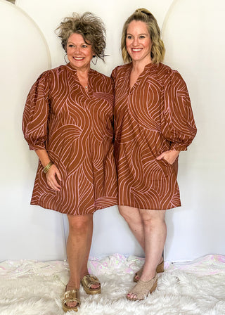 Two women modeling matching brown and rust-colored patterned mini dresses with puff sleeves, standing together against a white studio backdrop with a white faux-fur rug.