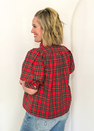 A woman with blonde hair tied back, wearing a red and green plaid flannel shirt and light blue jeans, standing against a white studio backdrop.