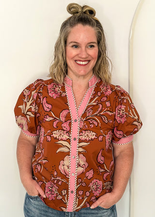 woman standing in a rust, pink and off white button up top and jeans.
