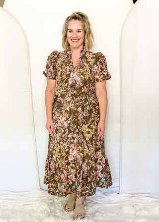 A woman stands in front of white arched backdrops wearing a brown floral midi dress with short puff sleeves and beige heeled sandals.