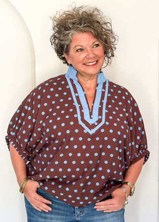 A smiling woman with short curly gray hair wearing a brown short-sleeve blouse with light blue polka dots and a light blue V-neck trim, paired with blue jeans.