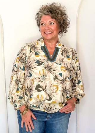A smiling woman with curly gray hair stands against a white arched wall, wearing a cream blouse with a large floral print in olive green, mustard yellow, and gray, paired with blue jeans.
