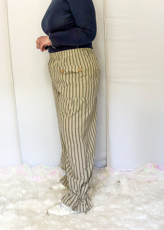 A person shown from the waist down wearing beige pinstriped cargo pants with a navy long-sleeve top and white sneakers, standing on a white faux fur rug in front of a white studio backdrop.