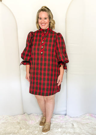A woman modeling a red and black plaid puff-sleeve dress with a high collar and front buttons, standing against a white arched backdrop.