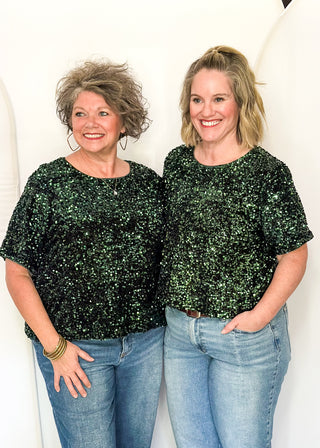 Two women standing side-by-side wearing matching dark green sequined short-sleeve tops and blue jeans, posing against a white arched backdrop.