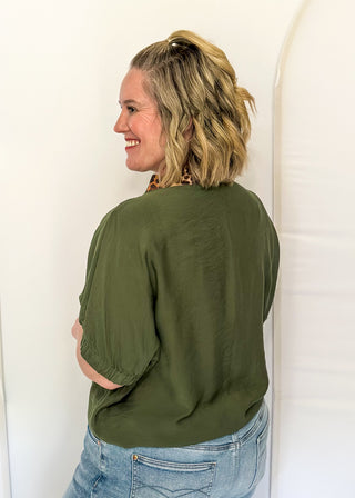 A woman with blonde hair tied back is shown from the back wearing an olive green short-sleeve top and light blue jeans, standing against a white arched backdrop.