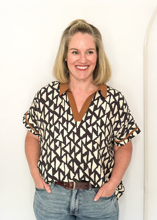 A smiling blonde woman wearing a short-sleeve patterned blouse with blue jeans, standing against a white arched studio backdrop.
