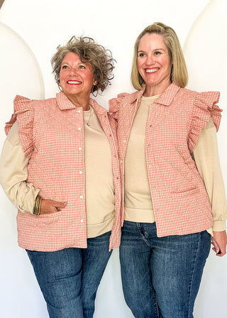 Two women smiling and modeling matching sleeveless button-front vests with ruffled shoulders in a pink and white gingham pattern, worn over beige long-sleeve shirts and blue jeans.