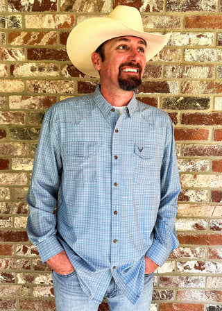 A man wearing a light blue and white checkered western button-down shirt, matching jeans, and a cream cowboy hat, standing against a rustic brick wall.