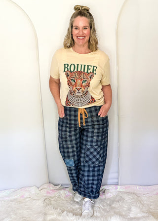 A woman modeling a beige graphic t-shirt with a leopard print and the word 'ROUIFFE' in green, paired with blue and gray plaid pajama pants, standing in front of a white arched backdrop.