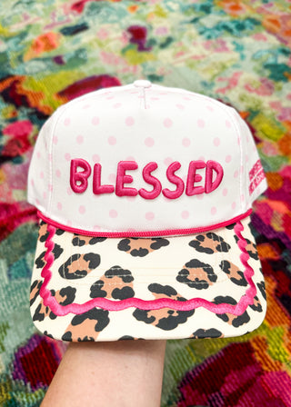A white snapback cap with pink polka dots, pink embroidered 'BLESSED' text on the front, and a leopard-print brim with pink trim, held up by a hand against a colorful patterned textile background.