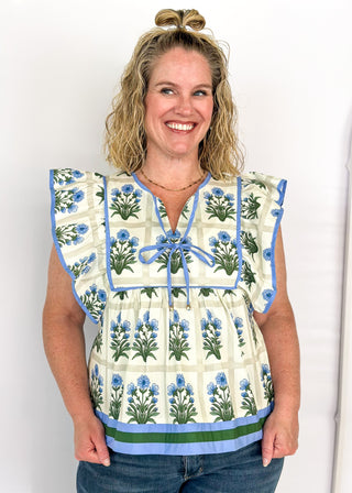 Flutter sleeve top with off white background and gray grid pattern-large blue florals with green stems/leaves in each grid.  Blue ribbon trim on sleeve and collar.  Blue ribbon tie neck.  Blue and dark green trim on bottom band. 