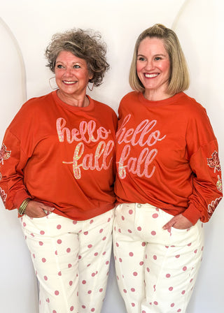 Two women wearing orange long sleeve tops with sequin leaves on the sleeves and "hello fall" across the front in sequins.