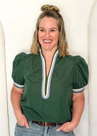 A smiling woman with blonde hair in a bun wears a dark green puff-sleeve blouse with a striped V-neck trim, paired with blue jeans, standing against a white arched backdrop.