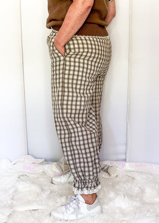A person wearing brown and beige gingham plaid pants, a brown sleeveless top, and white sneakers, standing against a white wall on a white faux fur rug.