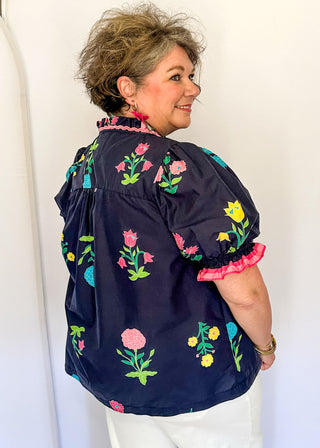 Navy floral short sleeve blouse with pink, green, yellow, and blue flowers, featuring puff sleeves, ruffled neckline, button front with pink trim, and a relaxed fit