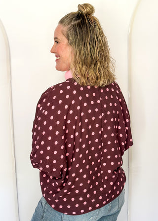 A woman shown from the back wearing a burgundy long-sleeve top with pink polka dots and light blue jeans, standing against a white arched wall.