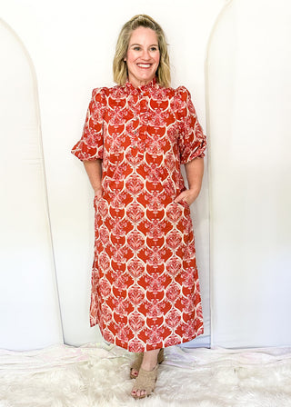 A woman stands barefoot in beige sandals against a white arched backdrop, wearing a short-sleeve midi dress with a red and white damask pattern and side pockets.
