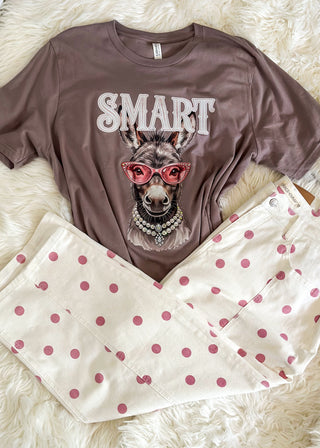 A flat-lay of a two-piece pajama set on a white faux fur background. The set includes a brown short-sleeve t-shirt with a graphic of a dog wearing pink sunglasses and the word 'SMART' in white, paired with cream-colored pants featuring pink polka dots.