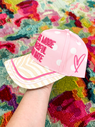 A pink baseball cap with white polka dots and pink embroidered text reading 'BE MORE POWERFUL AN HATE' on the front panel, held by a hand against a colorful patterned rug.