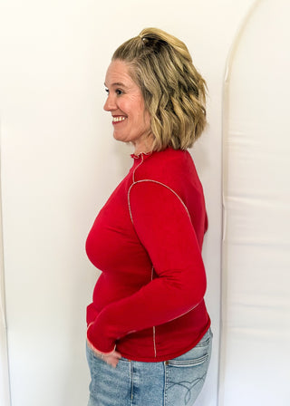 A woman with blonde hair tied back is shown in profile wearing a bright red mock-neck long-sleeve top with gold piping details and light-wash blue jeans, standing against a white studio backdrop.