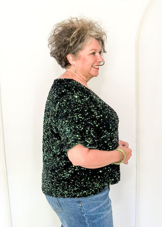 A woman with short curly gray hair is shown from the side, wearing a sparkling black and green sequined short-sleeve top and blue jeans, standing against a white arched studio backdrop.