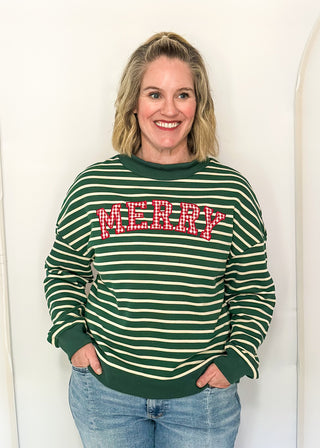 A woman wearing a green and white striped crewneck sweatshirt with 'MERRY' in red and white glitter letters across the chest, paired with blue jeans, standing against a white arched backdrop.