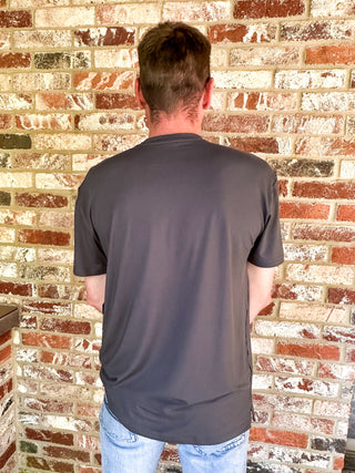 A man viewed from behind wearing a dark gray short-sleeve t-shirt and light blue jeans, standing in front of a red and white brick wall.