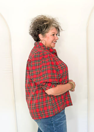 A woman with short, curly gray hair stands in profile wearing a red and black plaid flannel shirt and blue jeans, posing in front of a white studio backdrop.