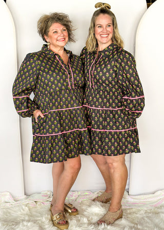Long sleeve black knee-length dress. Pink and green floral design. Tiered with pink piping detail. Buttons on top.
