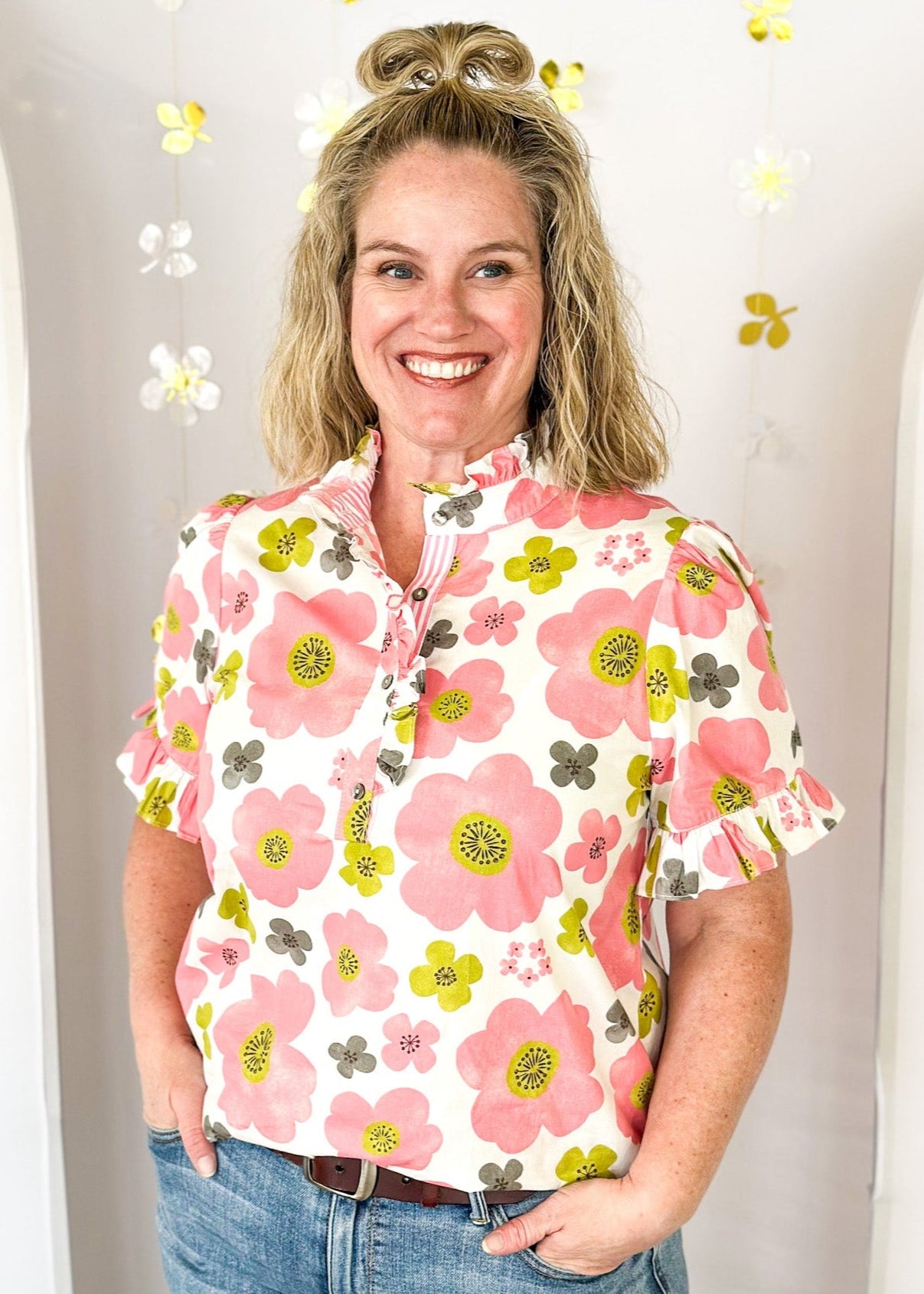 lightweight short sleeve blouse. Ruffle detail around the collar, sleeves and down the front highlighting the 4 button closure.  White background with distressed flowers in pink, lime and dark gray.