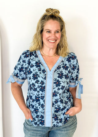 light blue top with navy flowers and red centers - light blue band around v-neck and straight down the front with blue and white striped trim along side it - light blue and striped bow at cuff