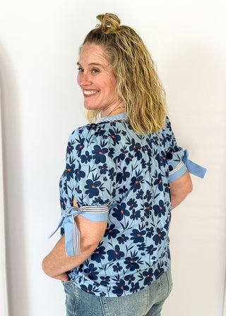 light blue top with navy flowers and red centers - light blue band around v-neck and straight down the front with blue and white striped trim along side it - light blue and striped bow at cuff